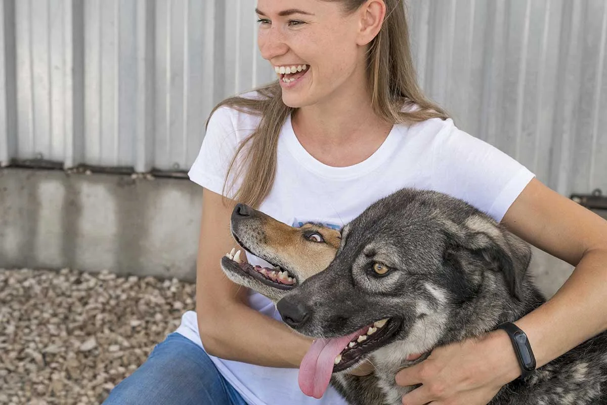 Smiling woman sitting outdoors happily hugging two medium-sized dogs, highlighting emotional support animal rights and housing considerations.